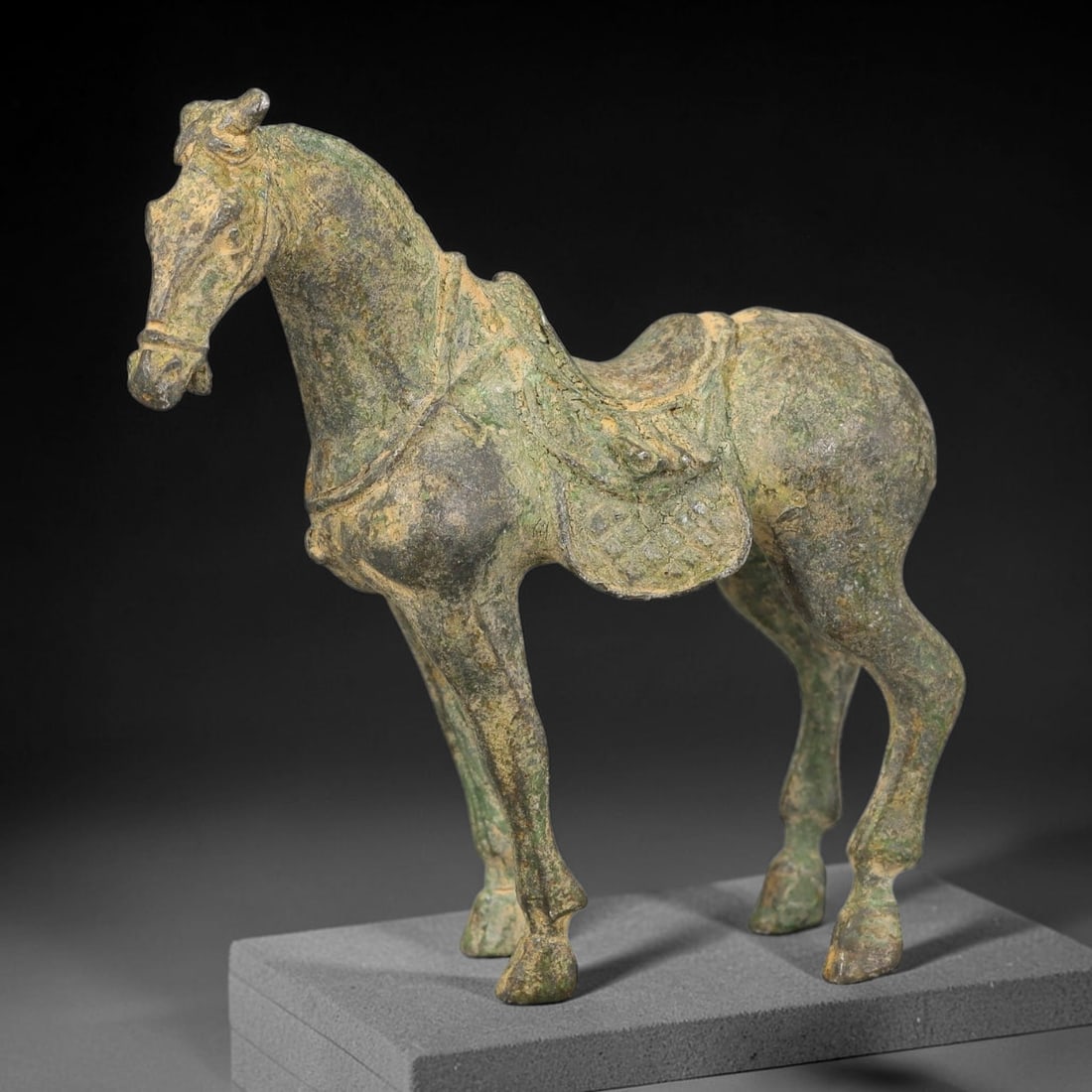A Chinese Bronze Standing Horse with Saddle Trappings, 76BT: Cast in bronze, the figure depicts a standing horse fitted with saddle and saddle trappings, with defined facial features, upright ears and slender legs. The surface bears encrusted green and brown pa