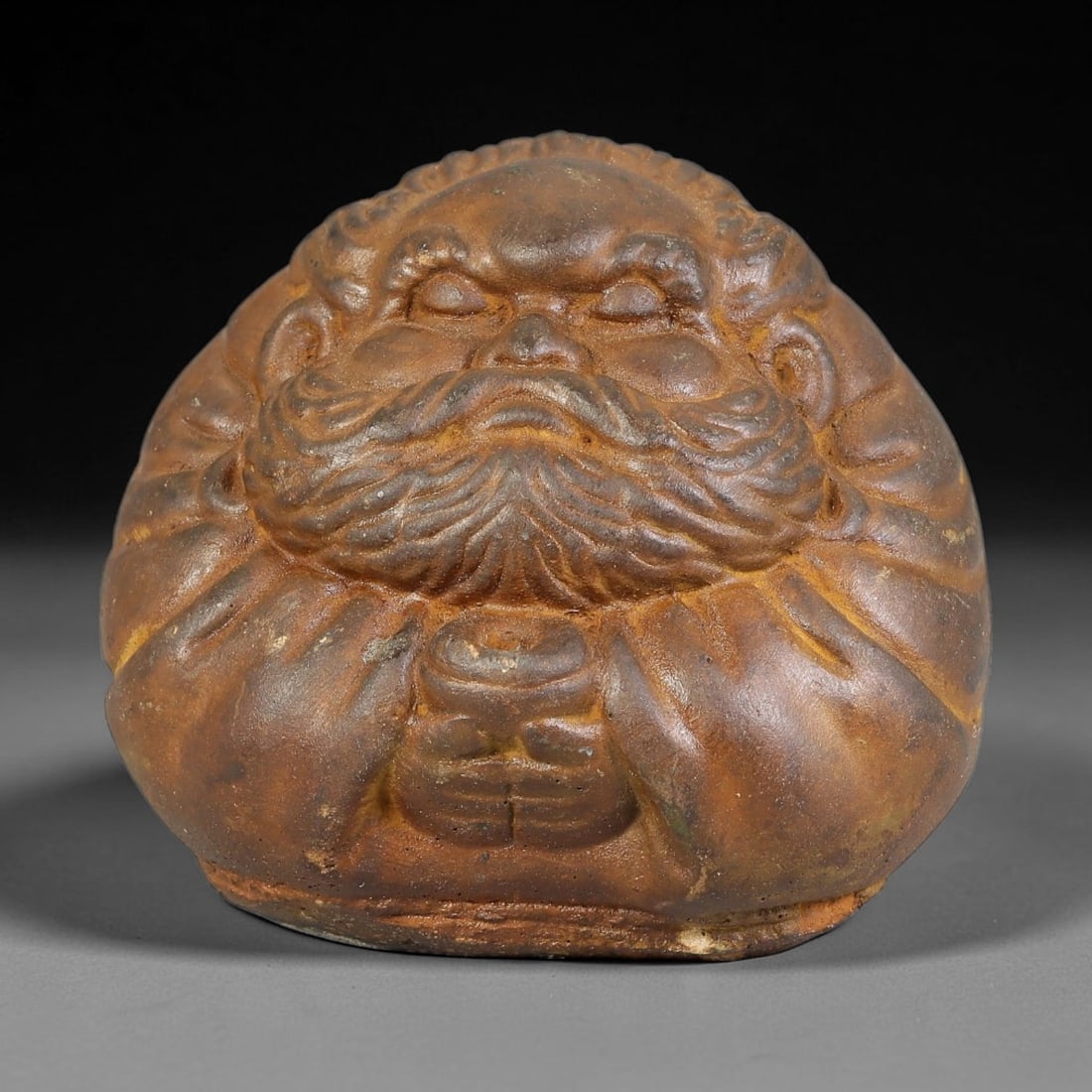 A Chinese Carved Stone Seated Damo Bodhidharma Figurine, 4BE: Carved from stone with rich aged brown patina and subtle surface wear, the figurine depicts a rounded seated Bodhidharma with closed eyes, thick curling eyebrows and full flowing beard. The figure hol