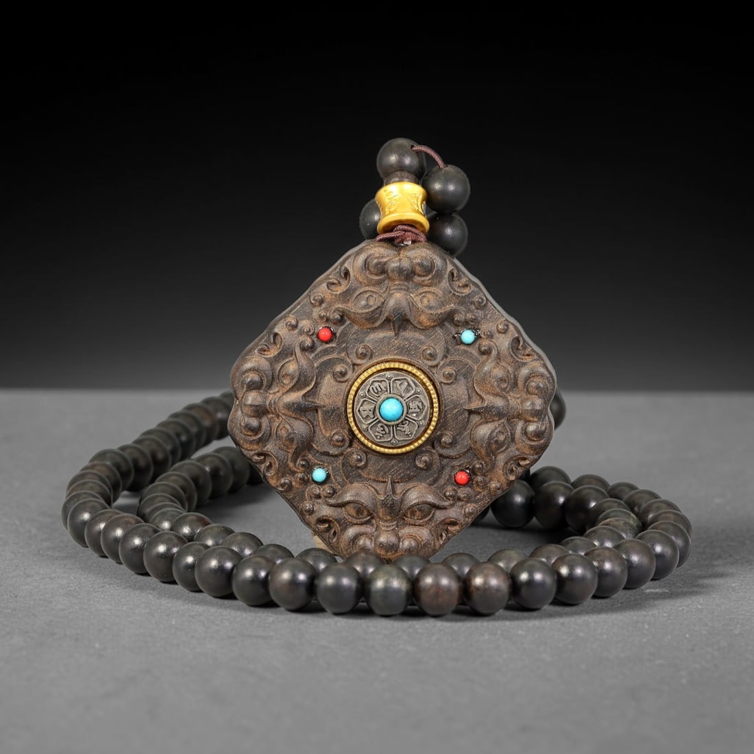 A Chinese Carved Wood Square Rotating Dharma Wheel Pendant with Beaded Necklace, 80BD: Square carved wood pendant with softly scalloped edges, deeply relief carved with paired beast masks across the face. The centre features a rotatable metal dharma wheel inlaid with turquoise coloured 