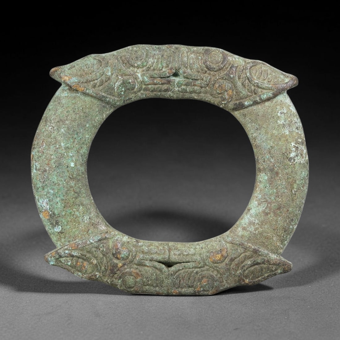 A Chinese Bronze Double Beast Head Ring Ornament, 71BT: This ancient bronze proto ring is cast with four stylized beast heads positioned around the circular band, each decorated with finely incised scroll and eye detailing. The bronze surface bears thick g