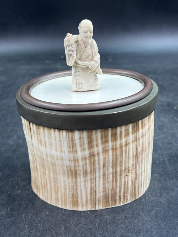 Japanese Okimono Figure on Brush Box: Figure depicts a man possibly a street vendor figure from folklore holding an object. Figure positioned on top of container or box with a ribbed or striated body and metal lid. Condition appears good.