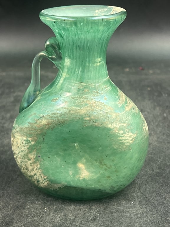 Antique Roman Glass Juglet: Likely dated between the 1st and 4th century CE. Pale turquoise or green blown glass with applied handle. Piriform (pear shaped) body, tooled indentations and surface weathering characteristic