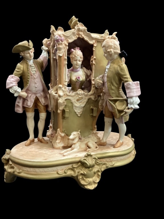 Royal Dux Porcelain Figural Group: Young woman in a sedan carriage with gentlemen . Dog resting nearby. Matte finish associated with Royal Dux. The grouping is 15 x 12 inches. - ()