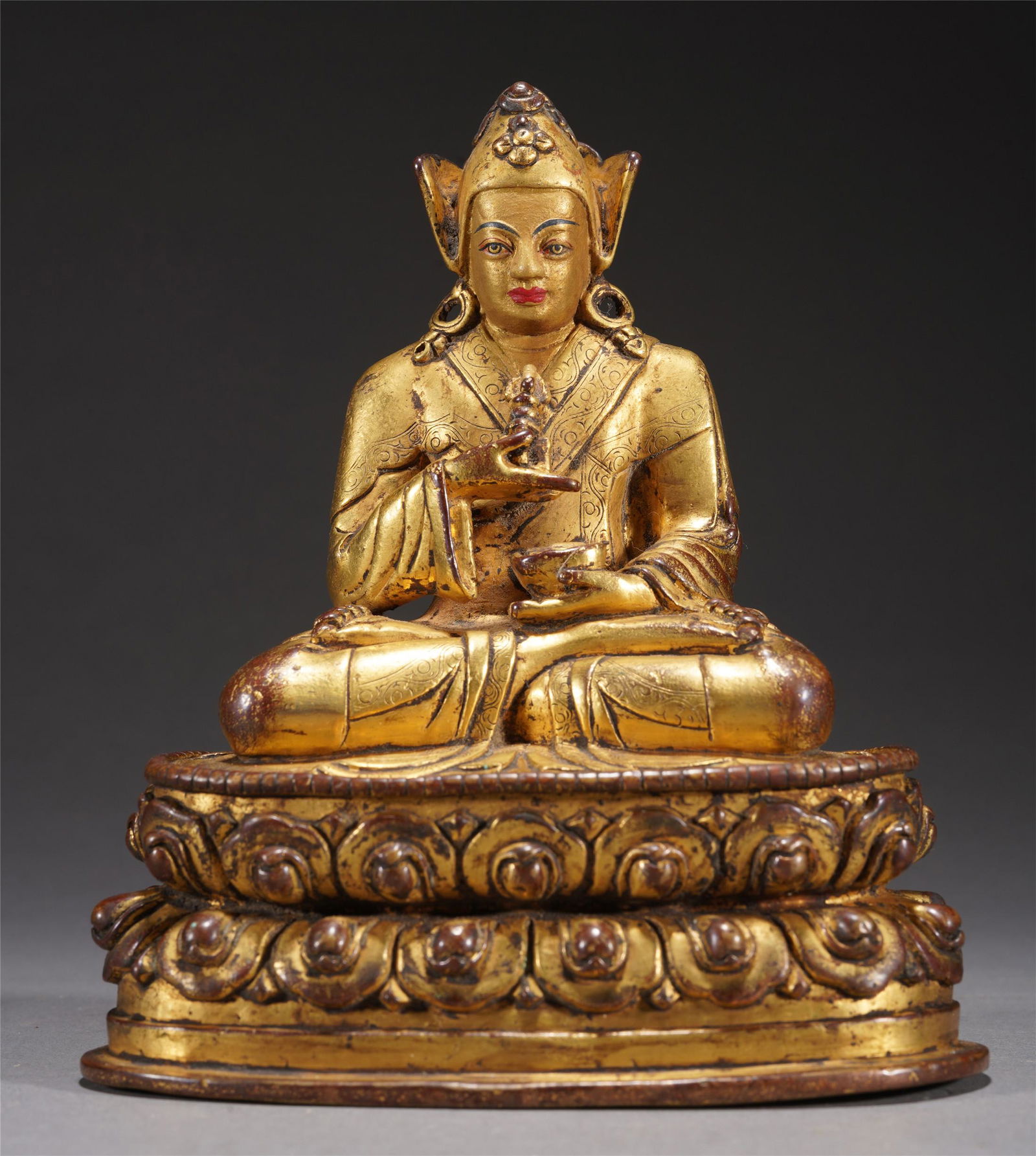 A TIBETAN BRONZE GILT PADMASAMBHAVA: A TIBETAN BRONZE GILT PADMASAMBHAVASeated on a lotus blossom holding a vajra in his raised right hand and a skull cup in his left, dressed in heavy brocade robes, the face with steady gaze surmounted