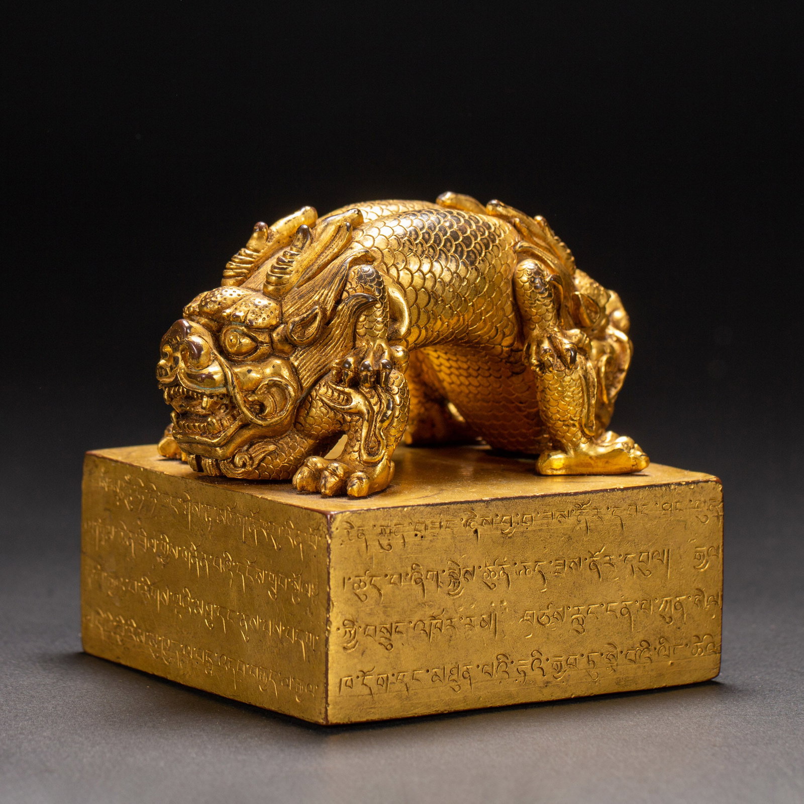 A TIBETAN BRONZE-GILT DRAGON SEAL: A TIBETAN BRONZE-GILT DRAGON SEAL The top carved with adorsed, crouching dragons flanked on either side, inscribed with tibetan unscript at the sides, richly gilt. Dimension: L. 8.6 cm. W. 8.6 cm. H. 