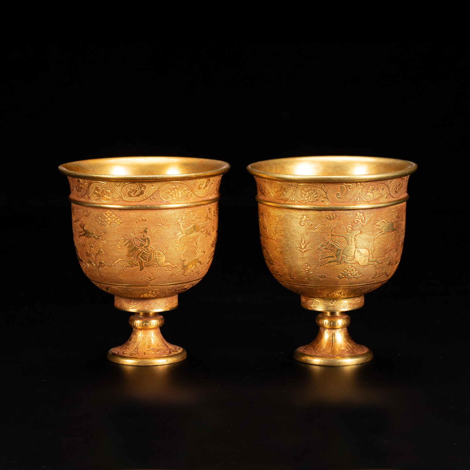 A Pair of Chinese Gilt Bronze Wine Cups: A Pair of Chinese Gilt Bronze Wine Cups A matching pair of finely crafted gilt bronze stem cups, each richly decorated with chased scenes of equestrian hunts amidst stylized foliage and animals. A con