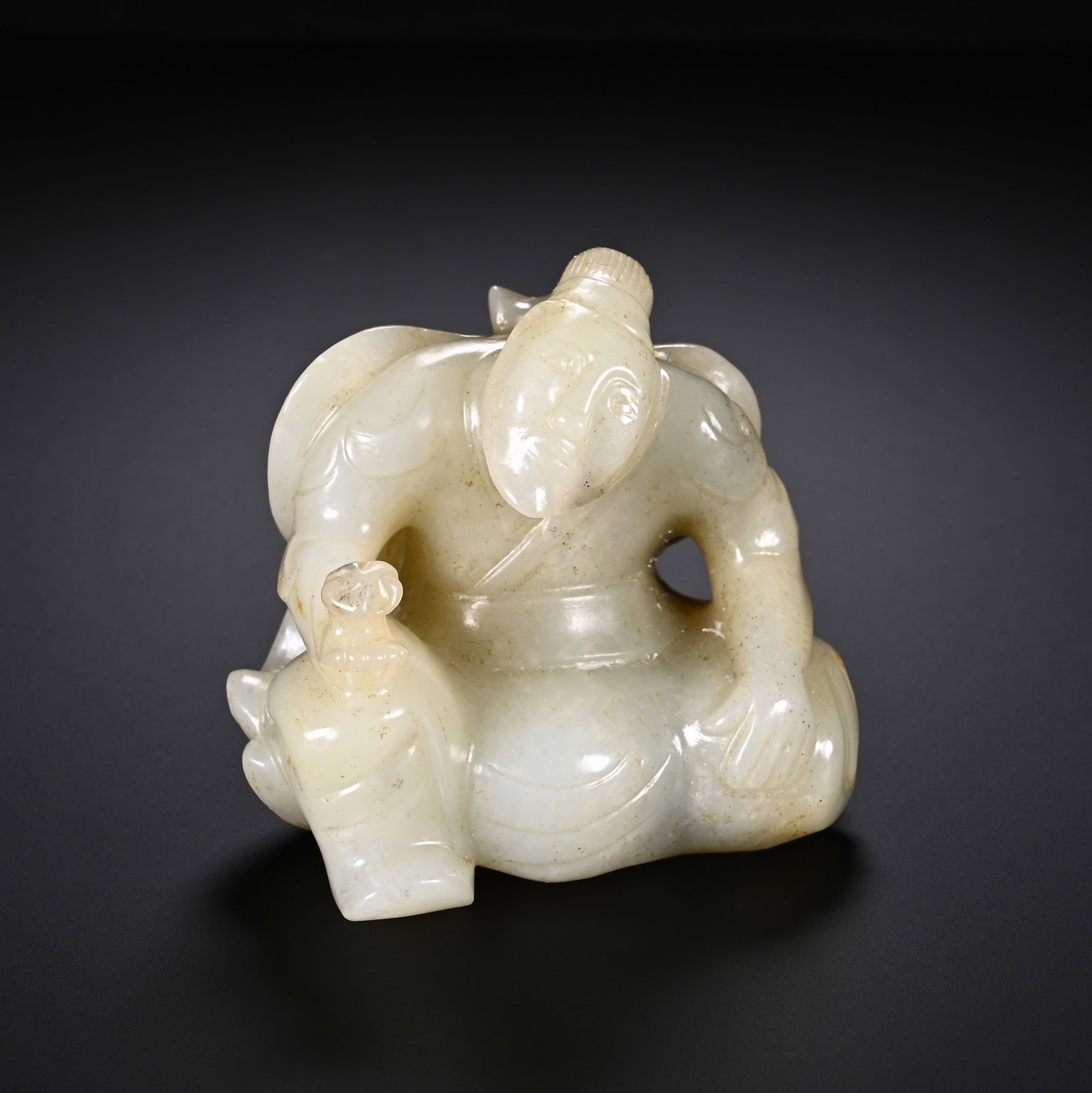 A Chinese White Hetian Jade Figure: A Chinese White Hetian Jade Figure A Chinese white Hetian jade carving of a seated figure, rendered in a relaxed pose with gentle facial features and flowing robes. The figure holds a small vessel,