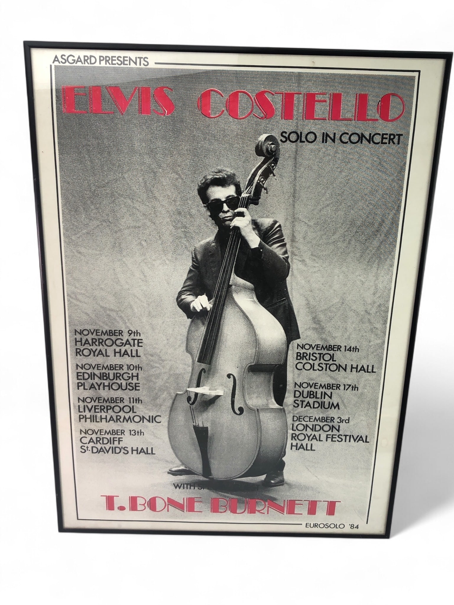 Elvis Costello Concert Poster Reprint: Framed reprint of an Elvis Costello 1984 concert poster featuring T. Bone Burnett. Measures 18 1/4"x 25 1/4"with frame. Includes tour dates and locations in the UK and Ireland. L5
