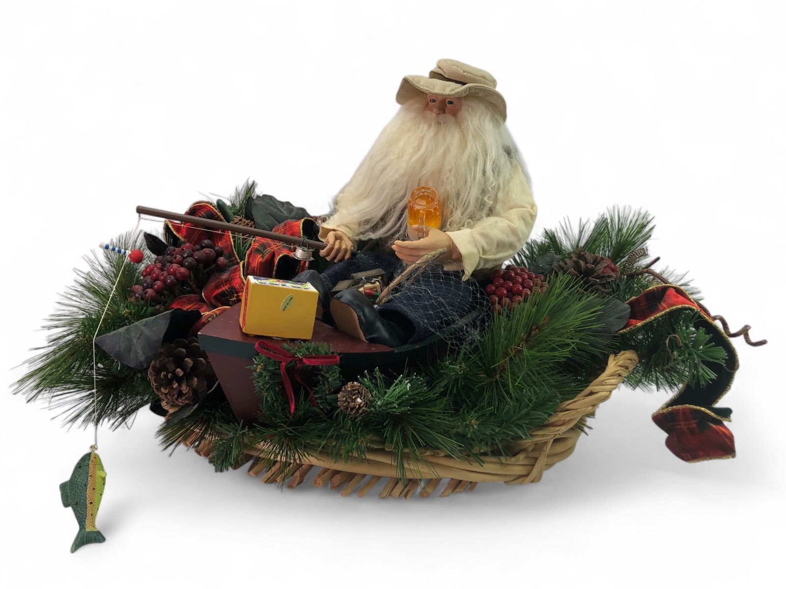 Christmas Fisherman Centerpiece Decoration: A 22" wide centerpiece featuring a fishing-themed Santa figurine in a boat surrounded by pine greenery, berries, plaid ribbon, and pinecones. The decoration is presented on a woven wicker base. Shows