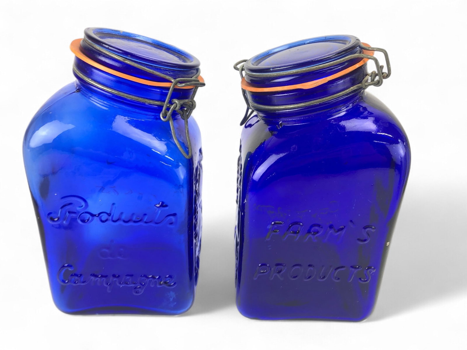 2 Casidis Milano Cobalt Glass Canisters: 2 Casidis Milano cobalt blue glass canisters featuring embossed fruit designs on the sides, tilted necks, and bail stopper closures with orange seals. Approximately 10 x 5 inches each.