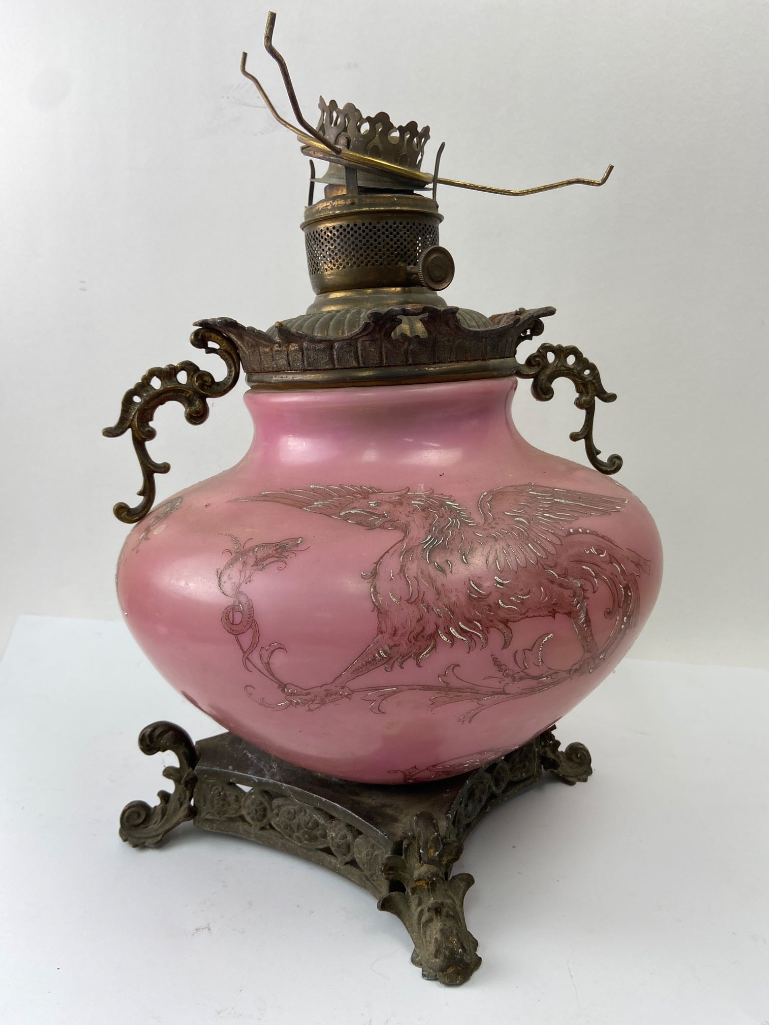 Pink Glass Oil Lamp With Griffin Design: Pink glass oil lamp featuring a detailed griffin design and ornate brass hardware. Measures approximately 13 inches wide. Not sturdy and does wobble. Includes a crown-like burner component and intrica