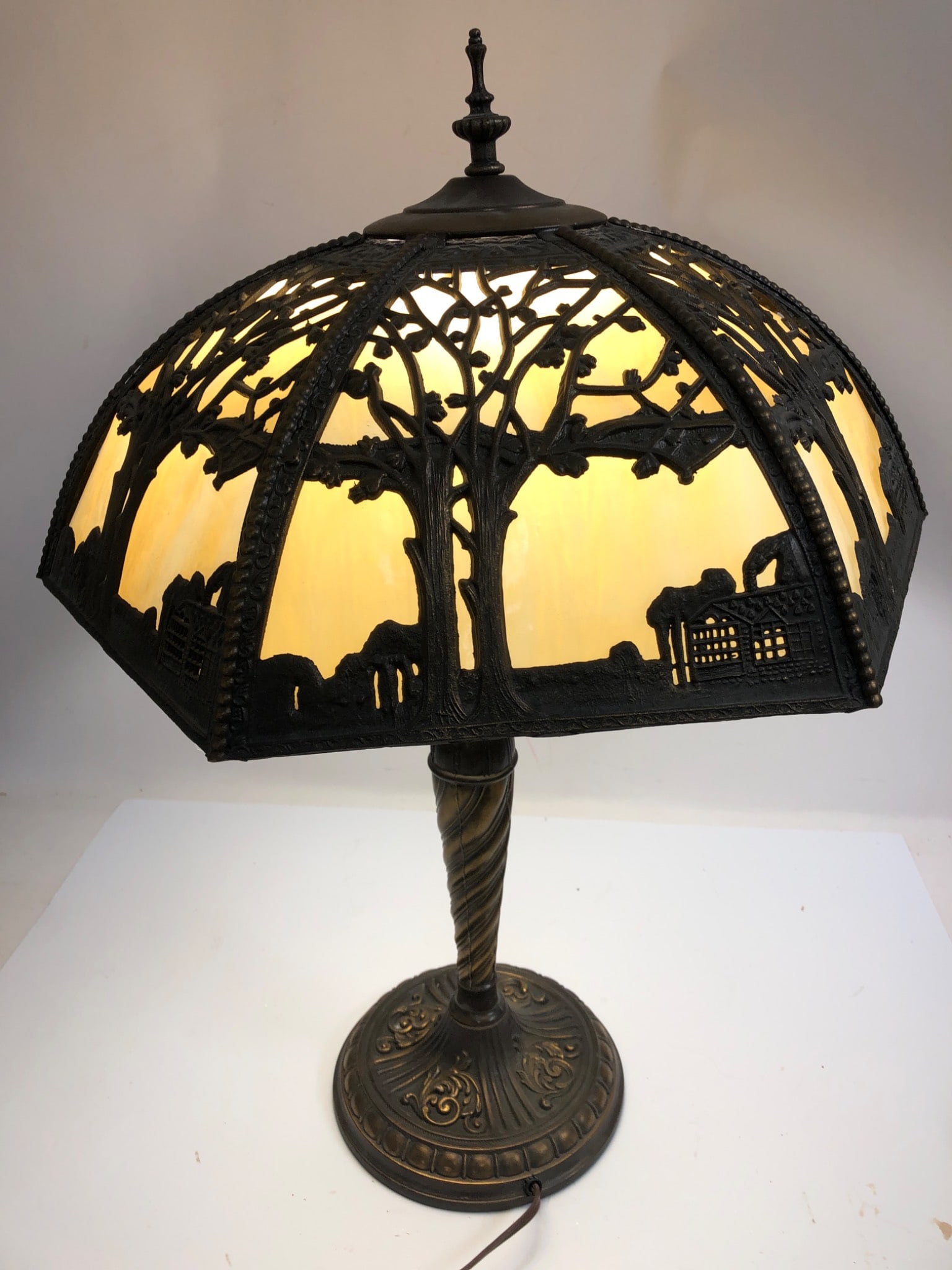 Slag Glass Table Lamp With Panel Shade: Metal table lamp standing 24 inches tall, featuring a detailed cast metal base and shade. The shade, 15 inches wide, showcases tree and landscape motifs, with yellow-toned glass panels. Electric wirin