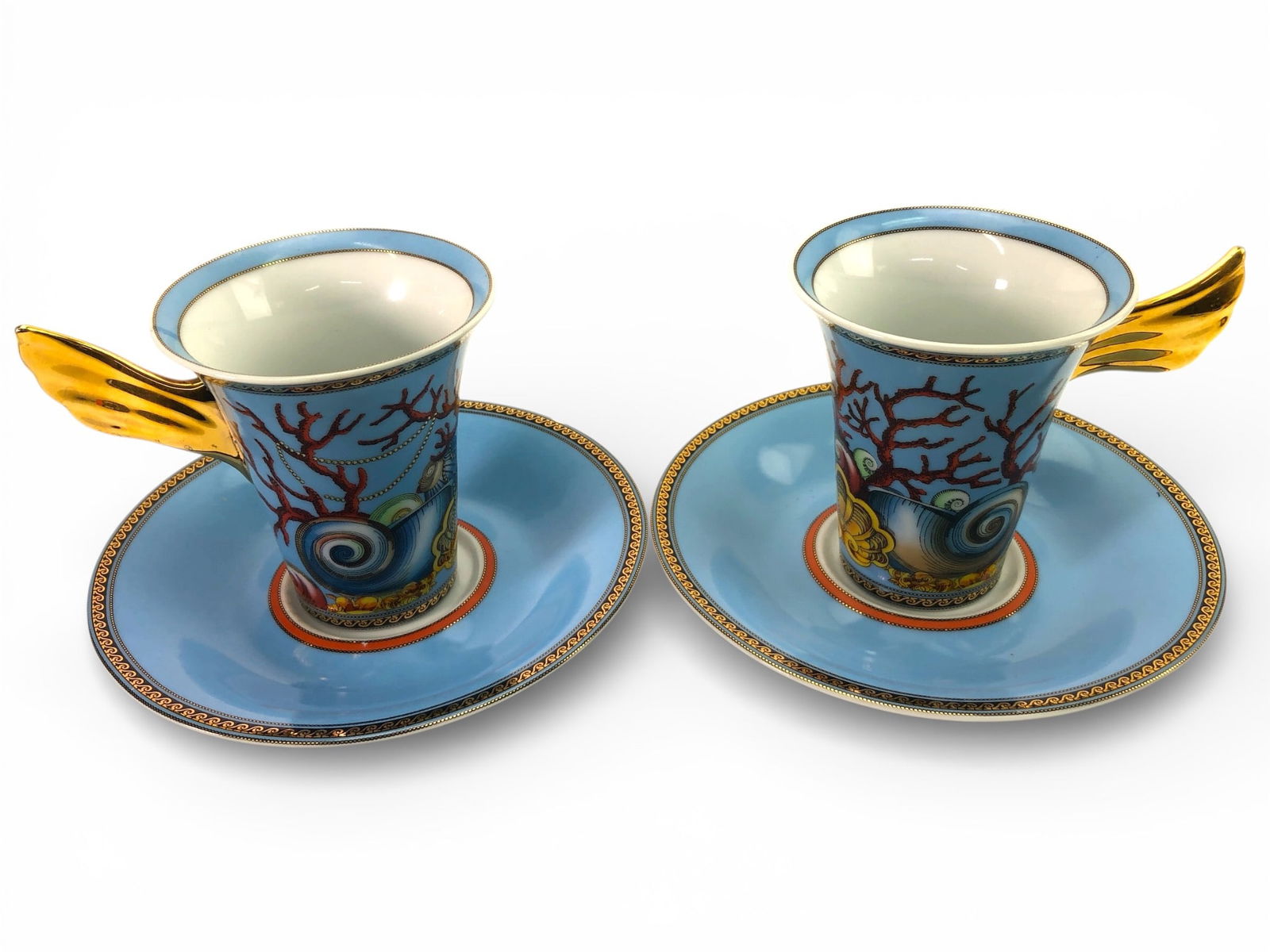 Versace Les Tresors De La Mer Cups & Saucers: Two Versace "Les Tresors de la Mer" porcelain cups with gold-tone wing-shaped handles & matching saucers. Saucer diameter: 5 7/8 inches; cup height: 3.5 inches. Features coral & shell motifs. Minor we