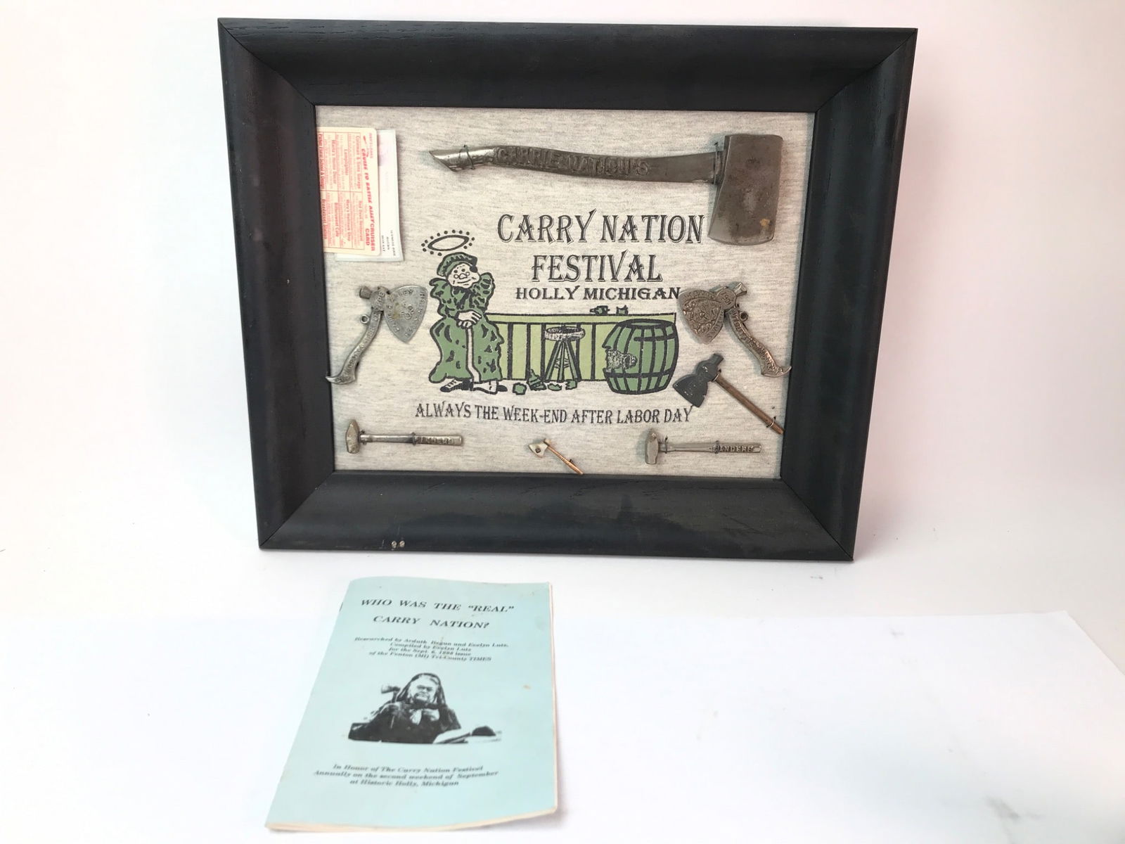 Carry Nation Festival Display With Artifacts: Framed display (17” x 14”) featuring items from the Carry Nation Festival in Holly, Michigan. Includes miniature axes, hammers, & printed memorabilia celebrating the event. Accompanied by