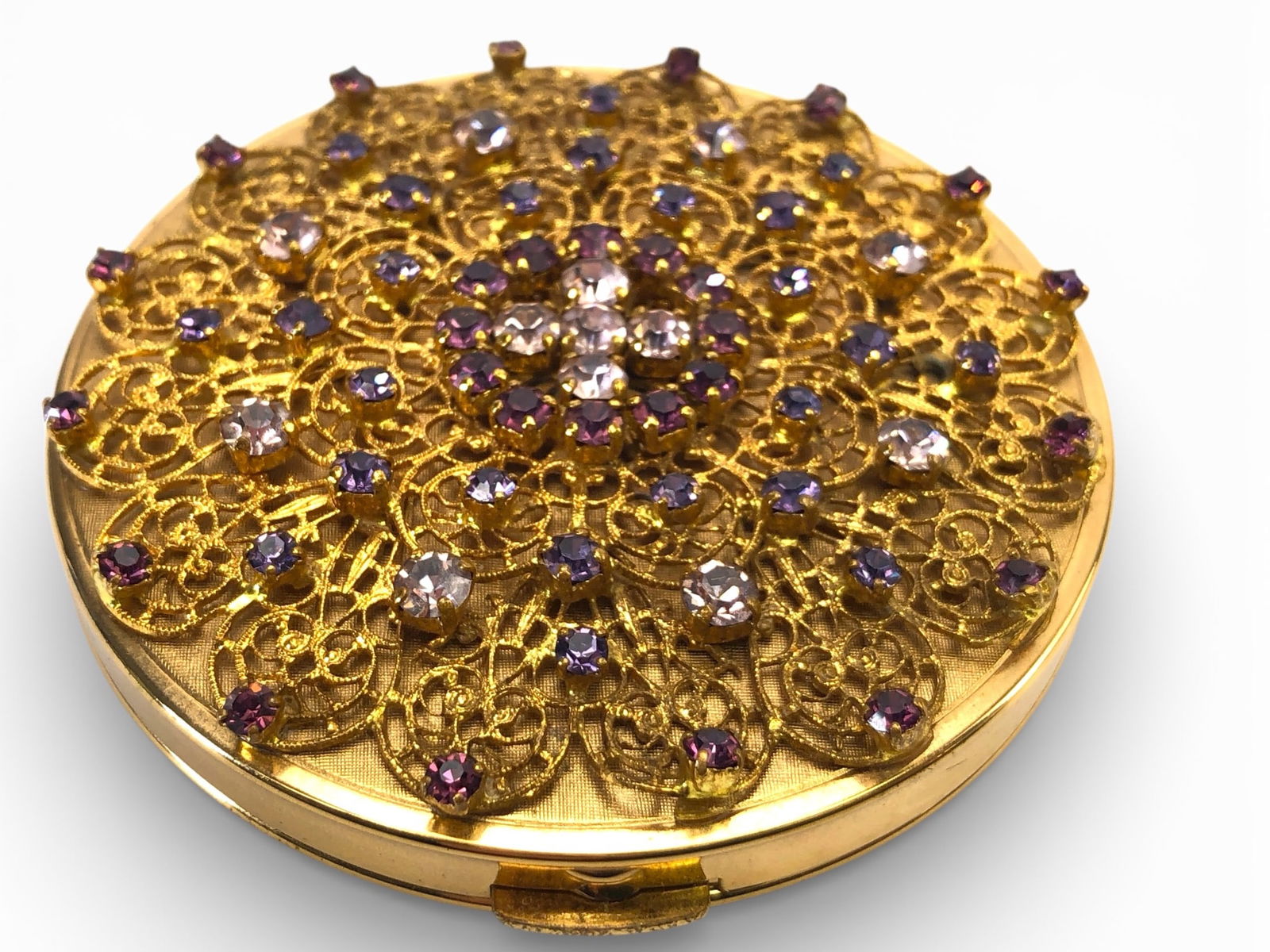 Vtg Germany Gold Compact W/Purple Stones: Vintage gold-tone compact measuring 2 3/4 inches in diameter, marked Germany. Features a filigree design adorned with purple and clear rhinestones on the lid. Interior includes a mirror, powder compar