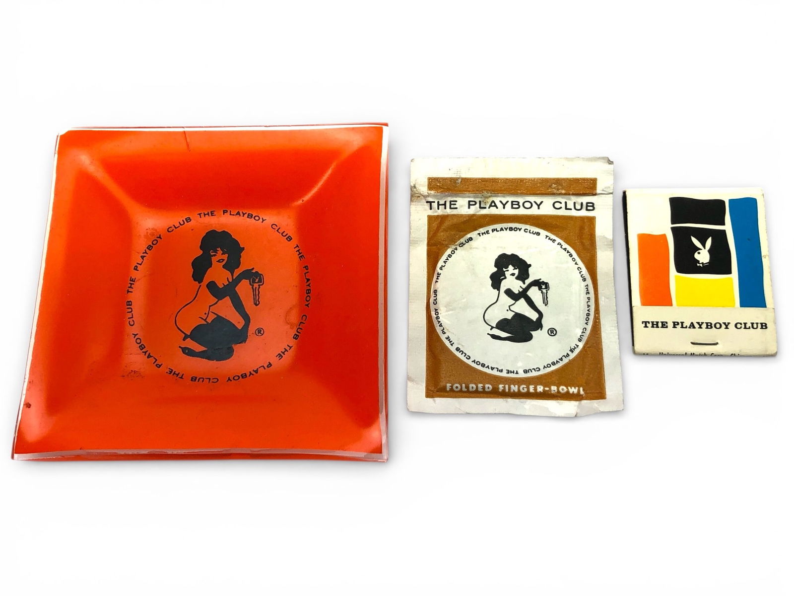 Playboy Club Dish, Matchbook & Towelette: Three Playboy Club memorabilia items: a square orange glass dish featuring the Playboy Club logo and imagery, a matchbook by Universal Match Corp with colorful design, and a 'Folded Finger-Bowl'