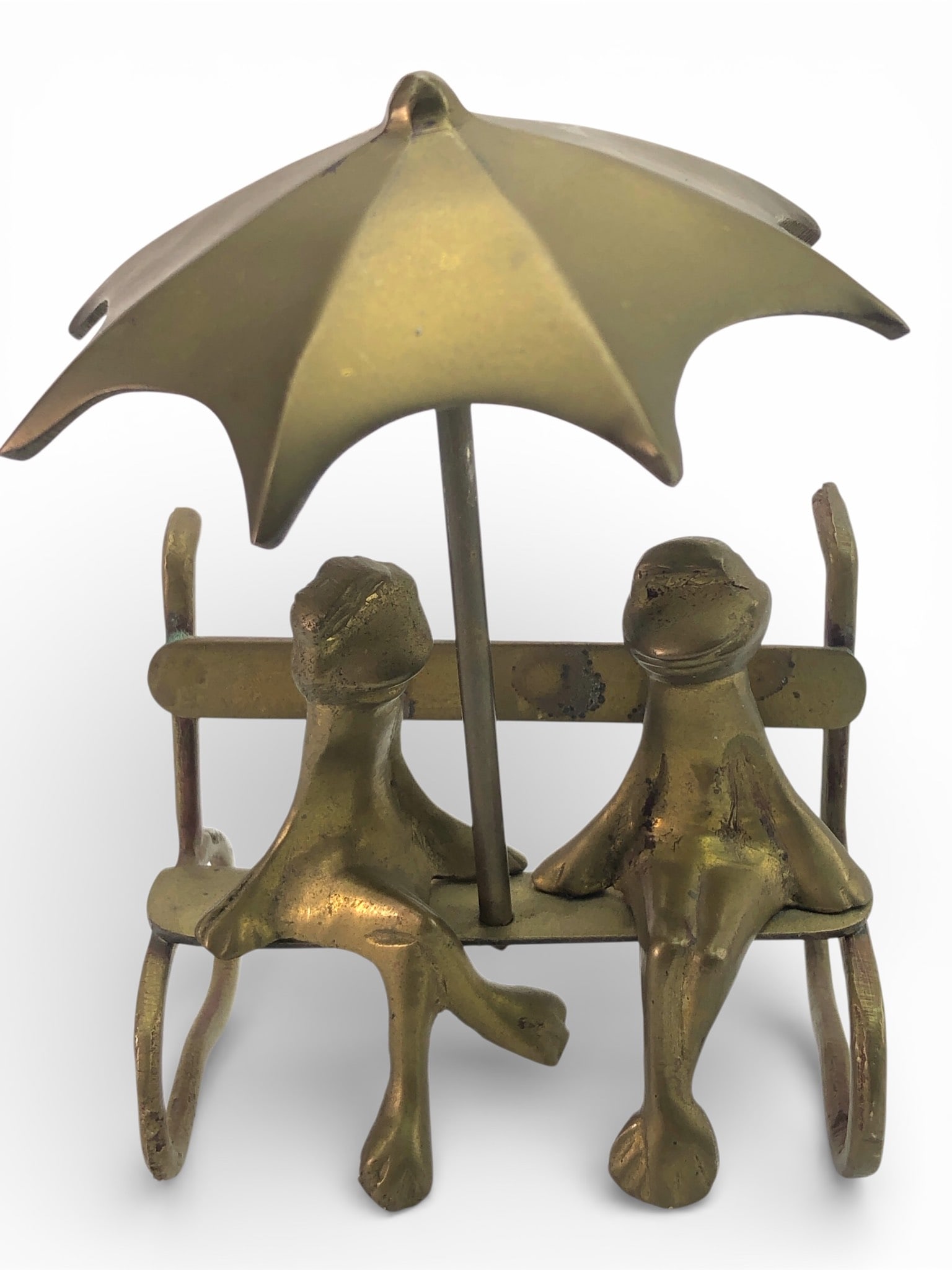 Brass Frogs On Bench Under Umbrella: Brass sculpture featuring two frogs seated on a bench under an umbrella. Measures 5.25 inches in height. Detailed craftsmanship with visible minor surface wear. The piece has a whimsical design and