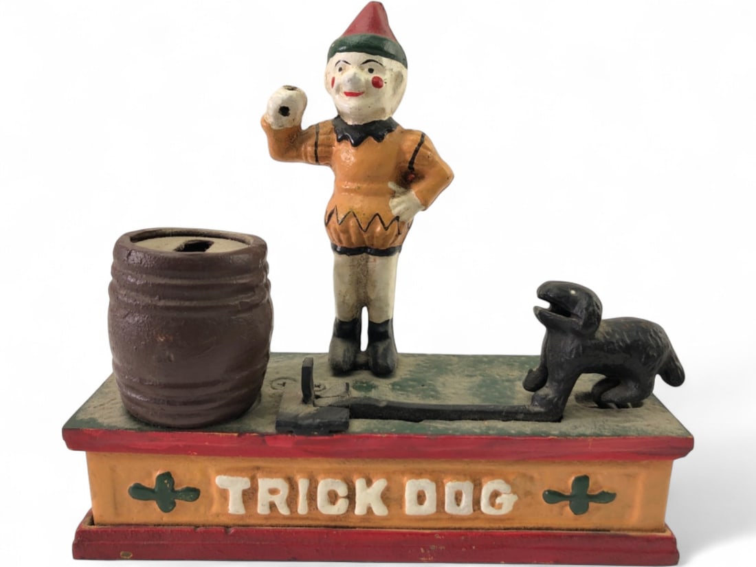 Cast Iron Trick Dog Mechanical Coin Bank: Cast iron mechanical coin bank depicting a clown and dog. Text on base reads 'TRICK DOG'. Coin slot on barrel. Mechanical action implied. Painted finish shows wear, chipping, and dust. Approx. 7" ht.