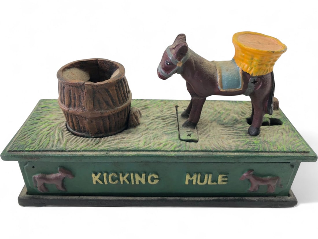 Antique Kicking Mule Mechanical Coin Bank: Cast iron mechanical coin bank featuring a kicking mule and barrel. Coin is inserted, and the mule kicks, depositing it into the bank. Text 'KICKING MULE' and 'BANK' on base. Decorative mule reliefs o