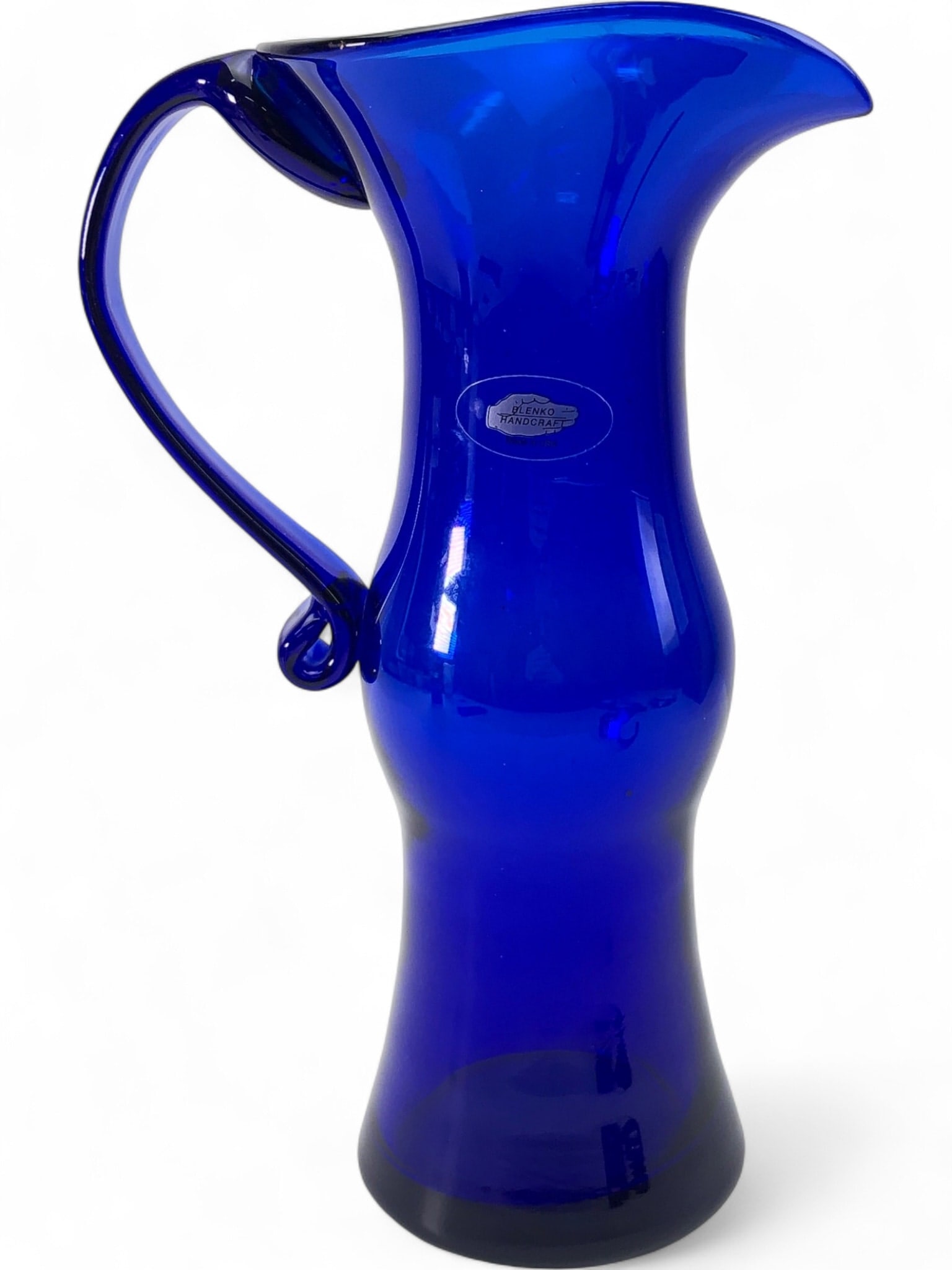 Blenko Handcraft Cobalt Blue Glass Pitcher: A hand-blown cobalt blue glass pitcher by Blenko Handcraft. This piece features a flared rim with a prominent pour spout, a cinched neck, and a bulbous body. The applied handle terminates in a decorat