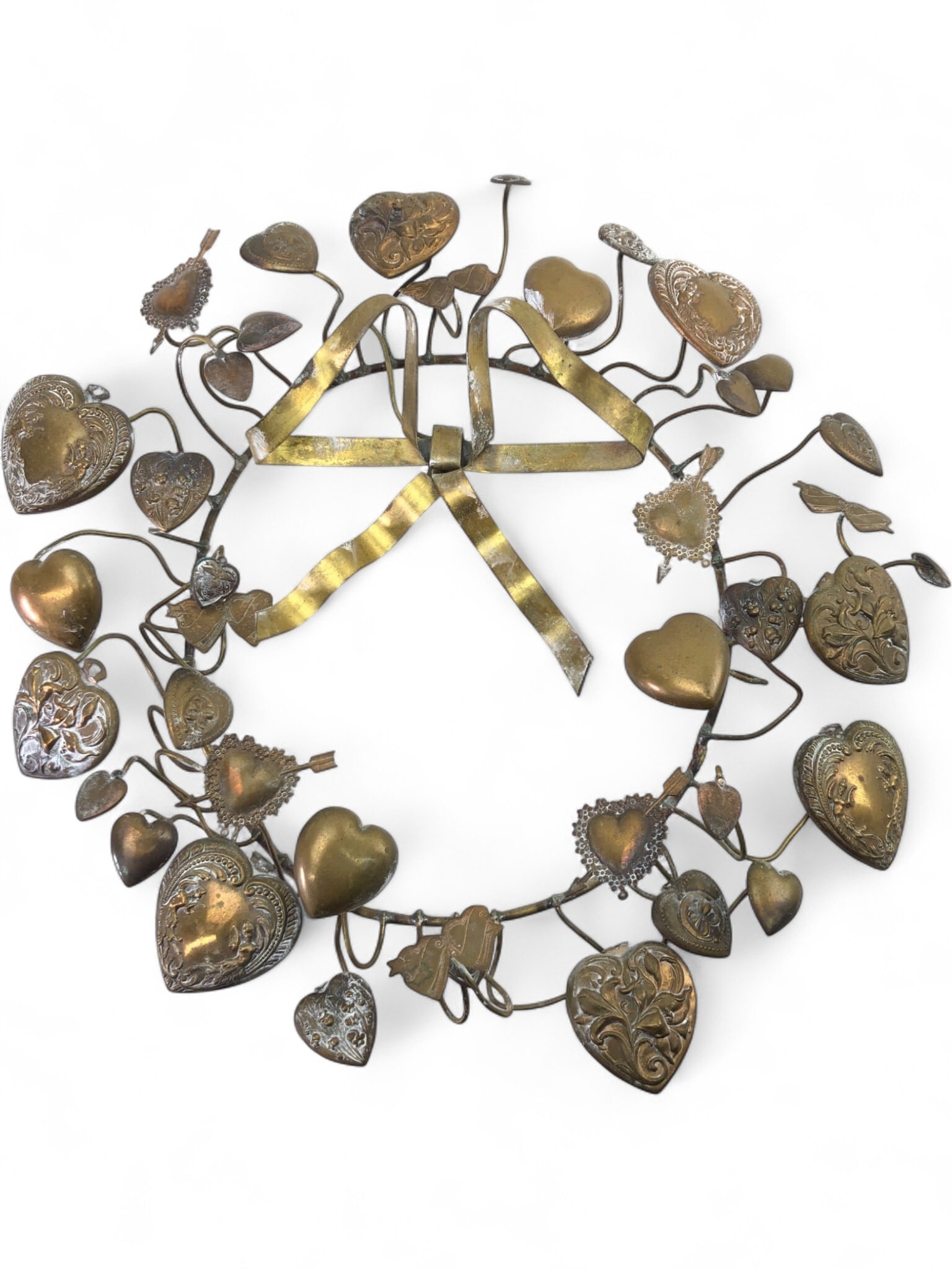 VTG Brass & Bronze Heart Wreath: A vintage decorative wreath crafted from brass and bronze-toned metals. Features a central bow and numerous hearts, some plain, others intricately embossed with floral and scroll motifs. Hearts vary i