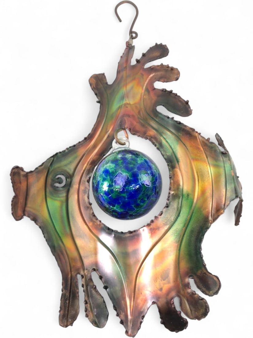 Hanging Copper Fish Sculpture with Glass Globe: Decorative hanging copper fish sculpture featuring a vibrant, iridescent paint in greens, oranges, and browns. A blue and green art glass globe, with a mottled appearance, is suspended within the fish