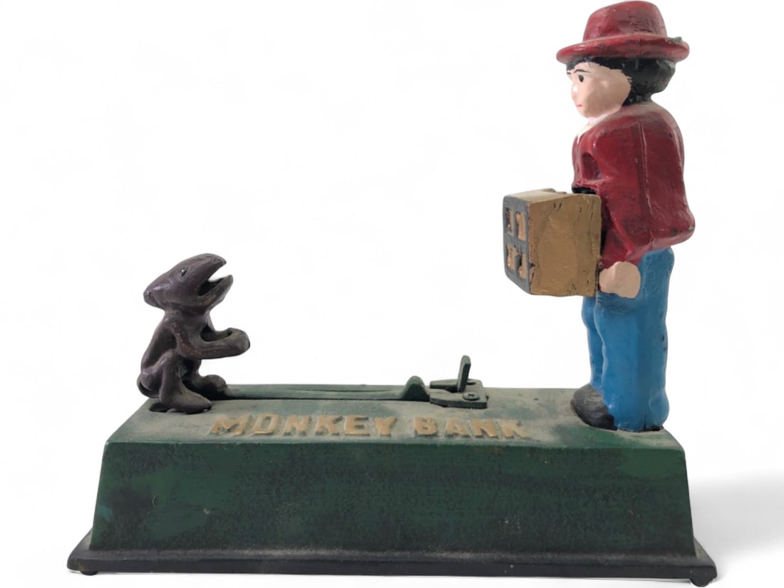 Cast Iron Monkey Mechanical Coin Bank: Cast iron mechanical coin bank, 7" tall. Features a man in a red hat and jacket holding a box, facing a monkey. The base is embossed "MONKEY BANK". Mechanical action involves the monkey's mouth. Paint