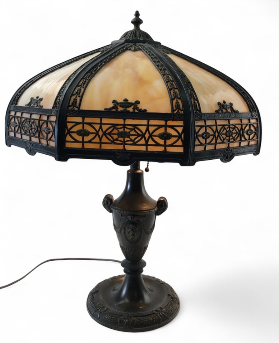 Bradley & Hubbard Style Slag Glass Table Lamp: An American Art Nouveau style table lamp featuring a six-panel caramel slag glass shade with ornate cast metal filigree overlay. The urn-form patinated metal base is decorated with ram's head