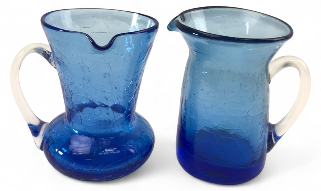 Pair Cobalt Blue Crackle Glass Pitchers 3.5" H: Pair of hand-blown cobalt blue crackle glass miniature pitchers with applied clear glass handles. One features a bulbous base and flared rim; the other has a tapered form. Both show rough pontil