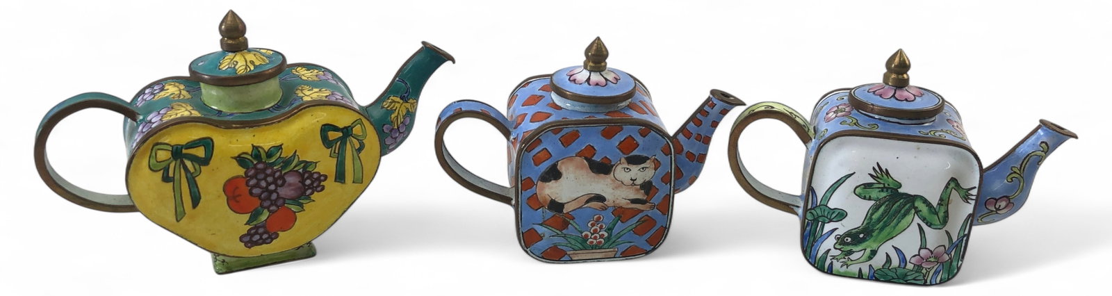 Set of Three Miniature Enamel Teapots: Set of three miniature painted enamel teapots. Unmarked. Features brass trim and finials with light blue speckled enamel interiors. Includes a heart-shaped yellow and green teapot with grapes, a