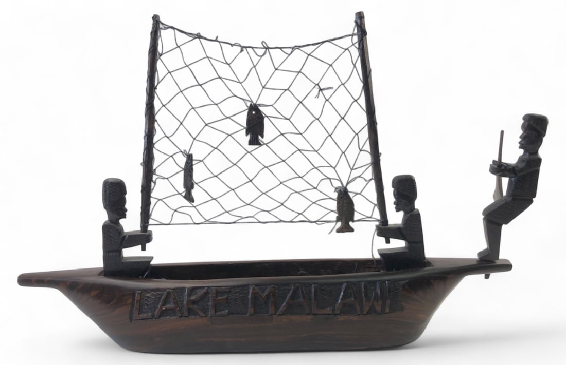 Kasimu Carved Wood Lake Malawi Fishing Boat 19": Carved wooden model of a fishing boat by Kasimu, originating from Mangochi, Lake Malawi. Features three removable carved figures, a string net with carved fish, and "LAKE MALAWI" carved on the side. T