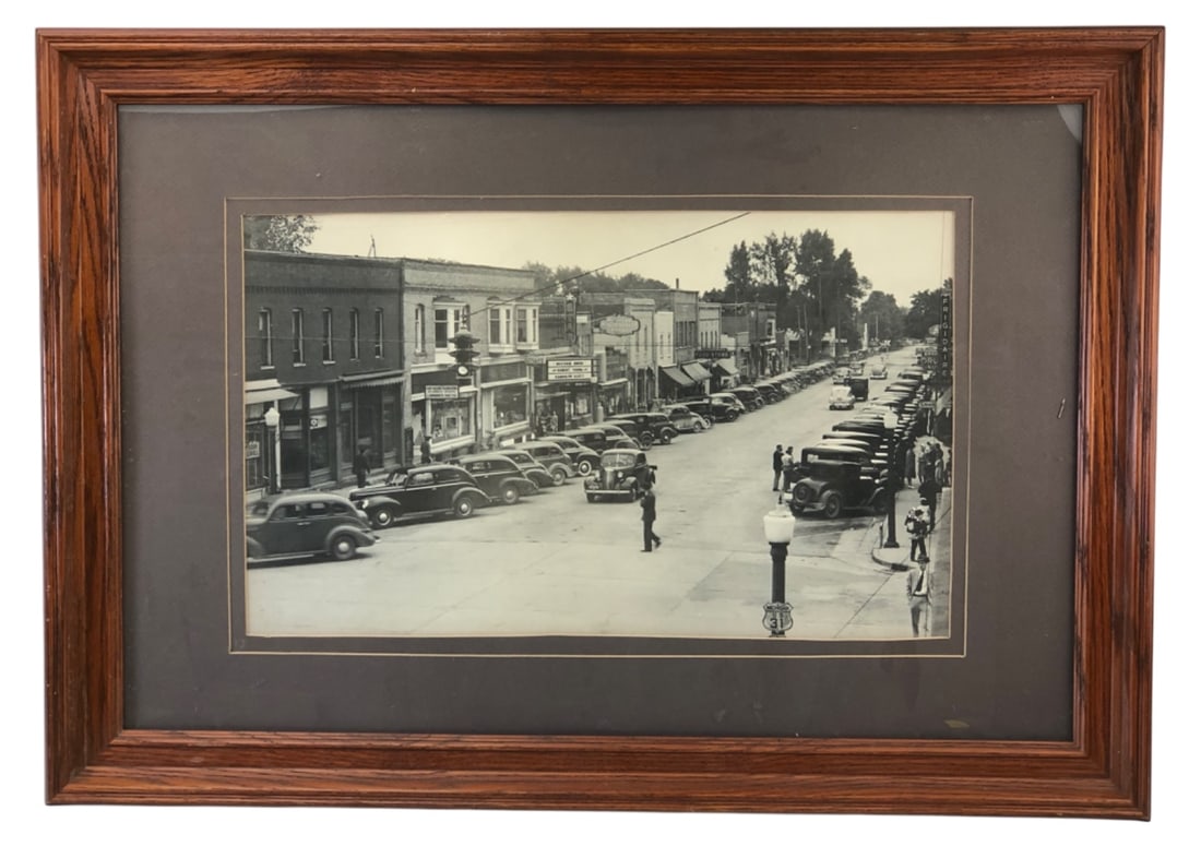 Framed 1941 Scottville MI Street Scene Photo: Framed black and white photograph of a 1941 street scene in Scottville, Michigan. Features diagonal parked vintage cars, storefronts, and a Michigan US 31 highway sign. Housed in a wooden frame with a
