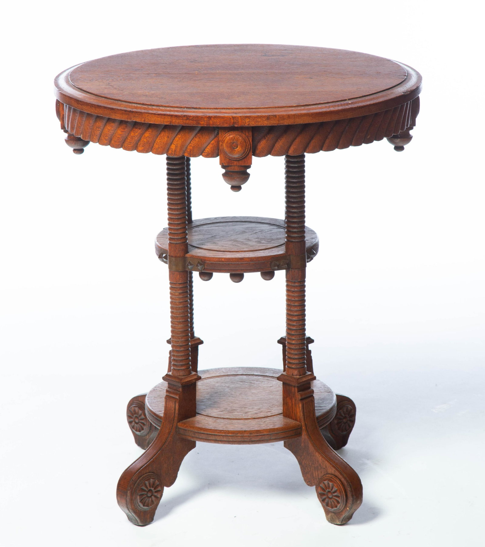 ROUND ARTS AND CRAFT PARLOR STAND.: American. Circa 1900. Oak with routed edge, base with turned and scrolled legs and applied rosettes, center shelf with brass decoration and lower shelf, 29"h. 24" diameter.