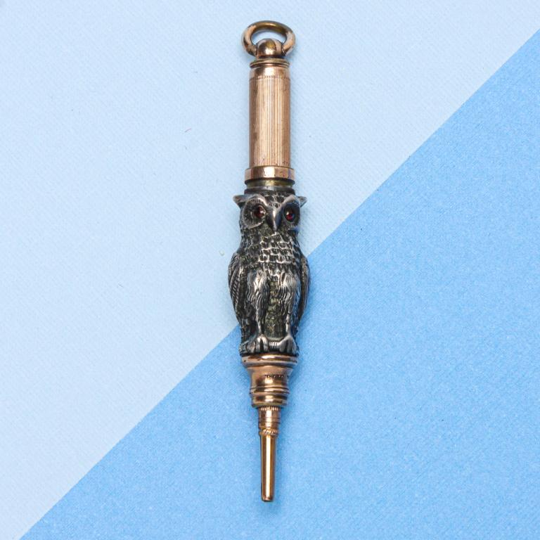 W.S. HICKS "MAGIC" OWL PENCIL.: Late 19th century. Novelty two-tone pencil made in the form of an owl sitting on a perch, with red glass eyes and attached suspension ring. Metal not tested, possibly sterling and low karat gold. Stam