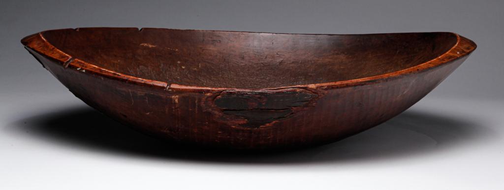 AMERICAN TRENCHER.: Nineteenth century. Oblong, rough hewn bowl with edge chips and age split. Split has make do repairs of tin and a brass escutcheon. 5.5"h. 26"l.