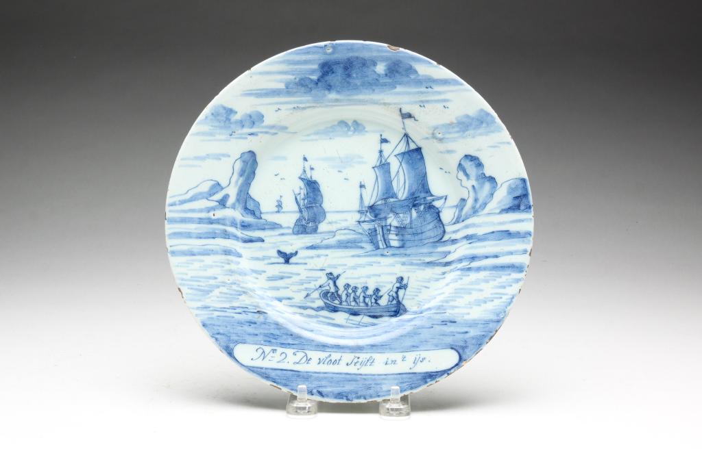 DELFT PLATE.: Late 18th century. Blue and white sailing ships with rowboat and whale "No. 2. De vloot Seylt int ys". Underglaze mark for The Porcelain Axe. Edge flakes, glaze crow's foot on the underside, light scr