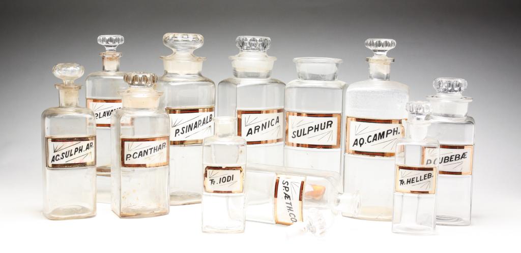 ELEVEN GLASS APOTHECARY JARS.: American, late 19th century. Whithill & Tatum reverse painted plaques on clear glass jars with stoppers. Cut and pressed glass stoppers. Including "Sulphur", "P. Canthar", "Arnica", "Sp. Lavand.Co", e
