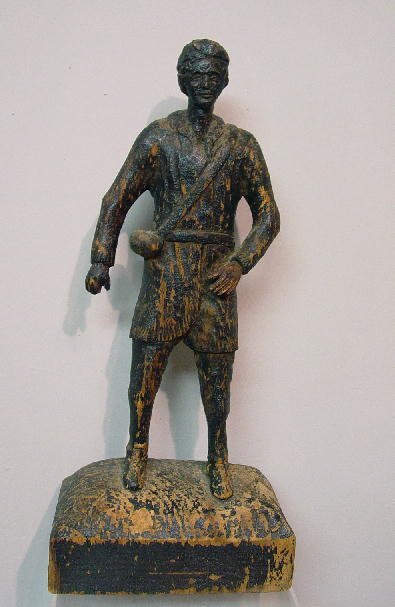 THREE FOLKY WOODEN CARVINGS. Frontiersma: THREE FOLKY WOODEN CARVINGS. Frontiersman, possibly Daniel Boone, wearing a fringed coat, powder horn and coonskin hat. Worn black paint. 13"h.; a seated dog with worn dark stain. Repairs. 6"h. And a