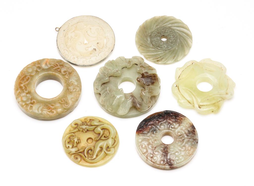 SEVEN ASIAN CARVED STONE DISKS.: Twentieth century or earlier. Various stones including jade. One pendant with white metal bezel, 2"d. and the rest bi or bi type disks with incised or relief designs including bats. 1.75" to 2"d.