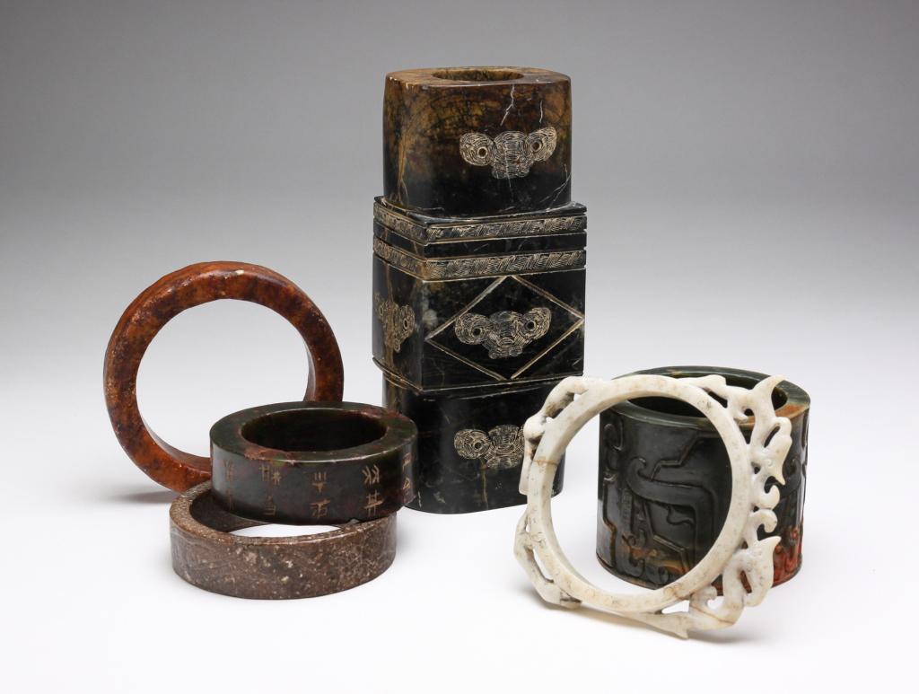 SIX CHINESE STONE CARVINGS.: Second half 20th century. Three heavy bangles, one with incised characters, one with relief carving, 3.25" to 4.5"d., ring with carved animals (split), 5.25"d. and a cylinder with relief masks, 3"h. a