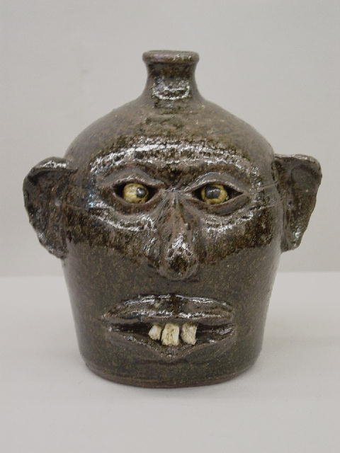 "LANIER MEADERS" GROTESQUE JUG. Dark green glaze.: "LANIER MEADERS" GROTESQUE JUG. Dark green glaze. Applied ears and strap handle with stone crossed eyes and teeth. Double incised lines around the eyes and around the head. 9"h.
