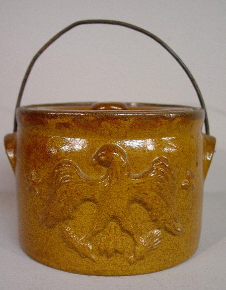 MOLDED POTTERY CROCK. Attributed to Kent, Ohio. Ye: MOLDED POTTERY CROCK. Attributed to Kent, Ohio. Yellow clay with a molded American eagle and two stars on one side. Grainy dark amber glaze. With lid and wire bale handle. 4 3/4"d. 3 1/2"h.