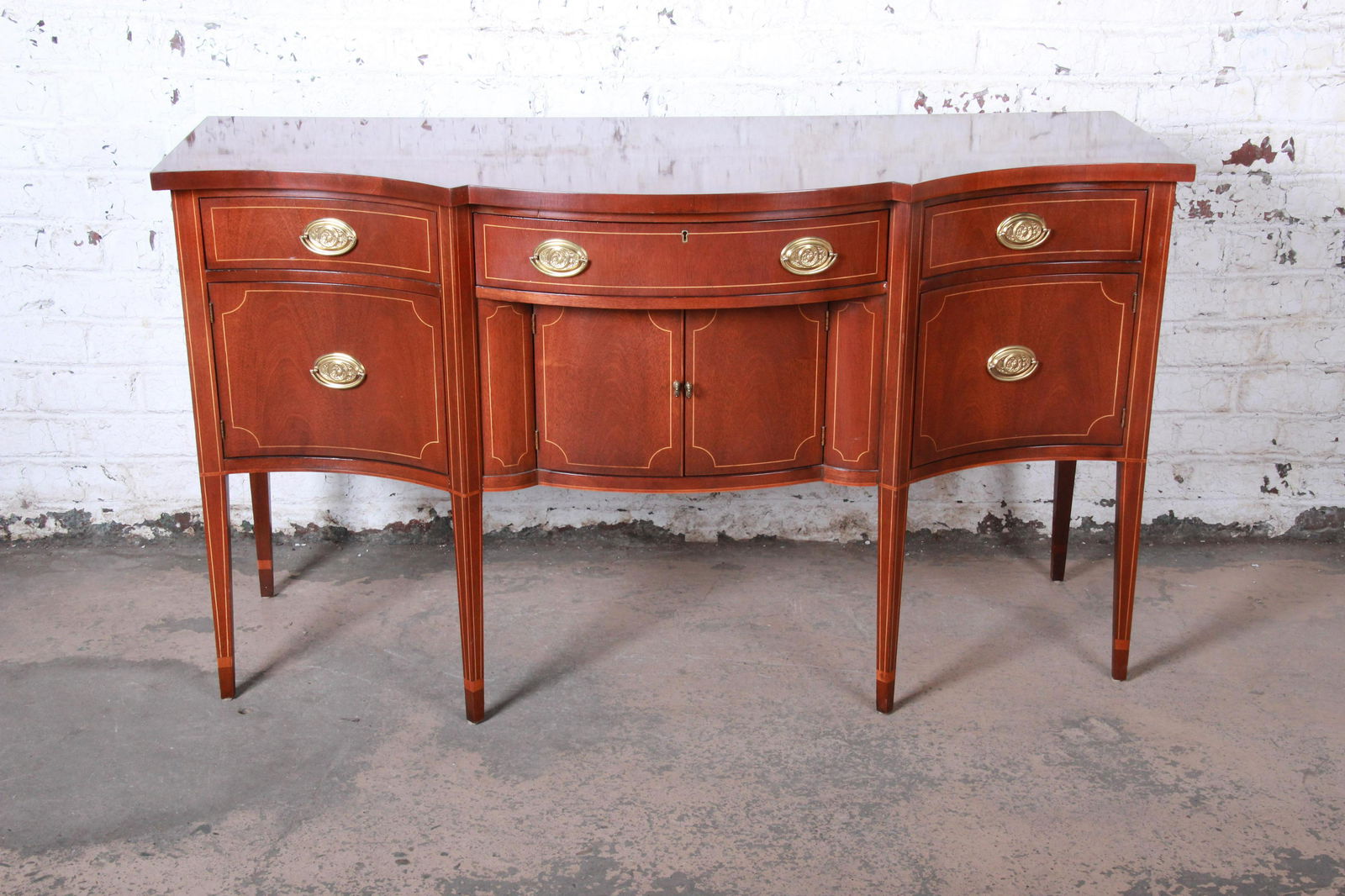 Baker Furniture Hepplewhite Style Inlaid Mahogany Bow: A gorgeous Hepplewhite style mahogany sideboard buffet by Baker Furniture. The sideboard features stunning mahogany wood grain with string inlay and a nice bow front design. The case rests on tall tap
