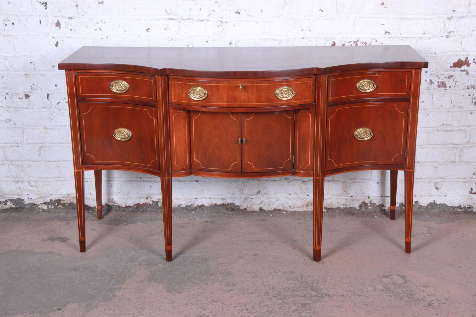 Baker Furniture Hepplewhite Style Inlaid Mahogany Bow: A gorgeous Hepplewhite style mahogany sideboard buffet by Baker Furniture. The sideboard features stunning mahogany wood grain with string inlay and a nice bow front design. The case rests on tall tap