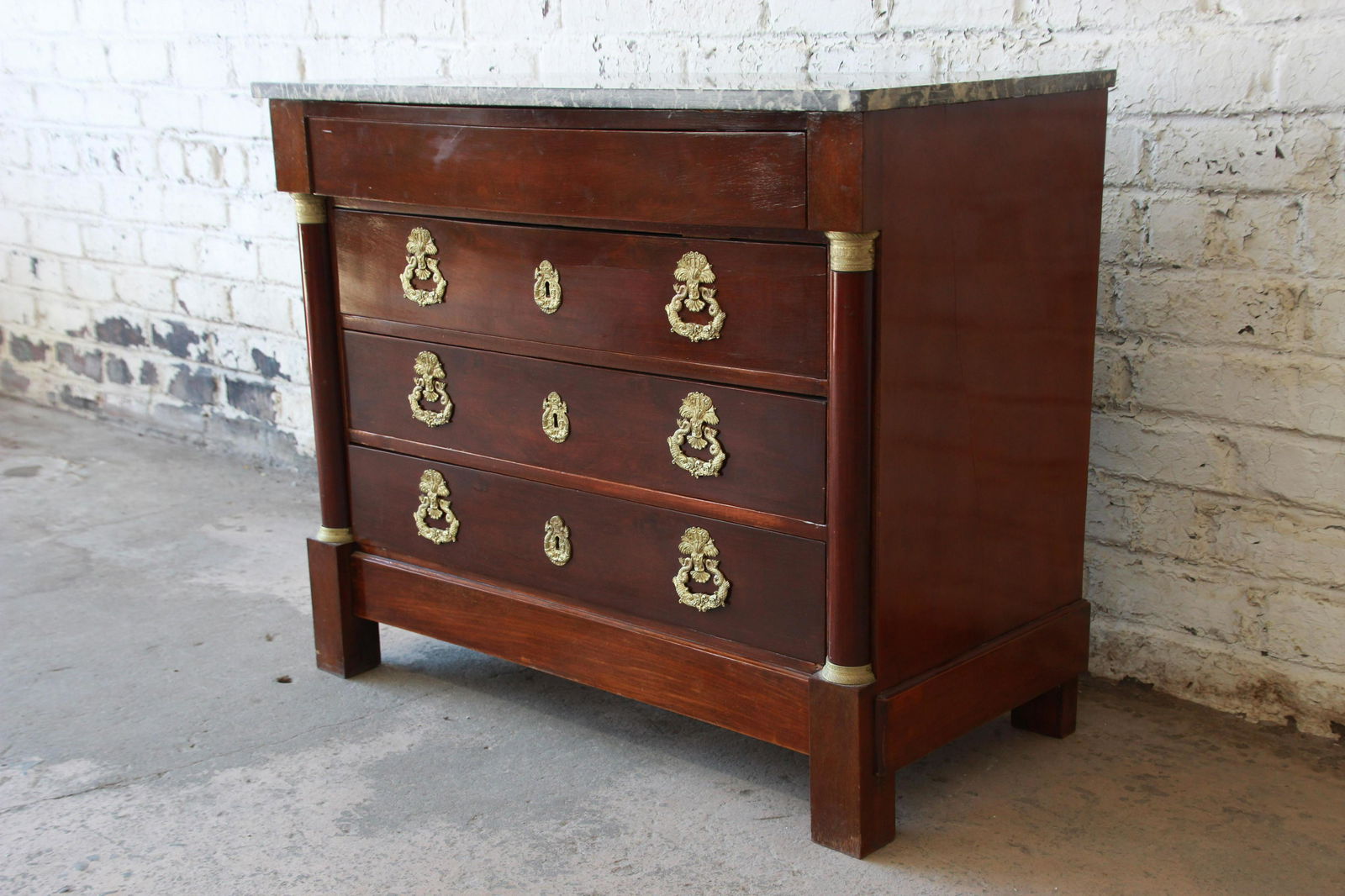 French Empire Mahogany Marble Top Commode Chest of: Offering a beautiful mahogany French Empire commode chest of drawers from the 1850s. The four drawer chest has unique ornate brass hardware and brass accents. It has a gray marble top complimenting th