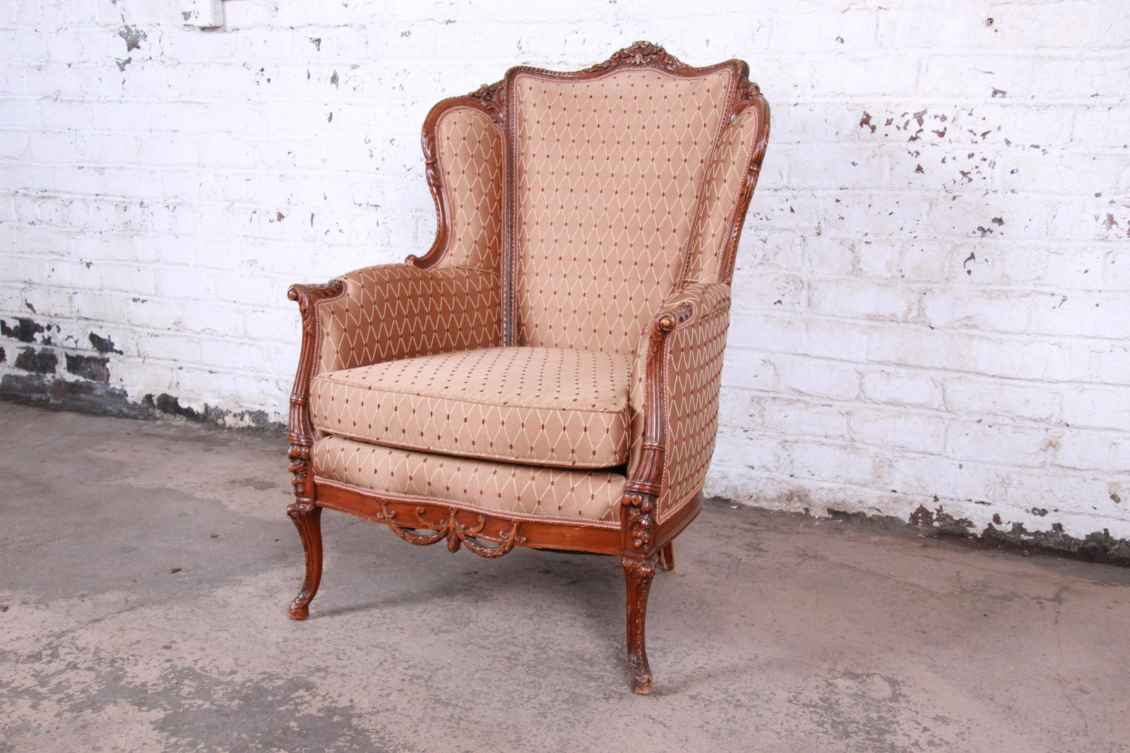 Antique French Carved Wing Back Lounge Chair: A gorgeous antique French carved wingback lounge chair circa 1930s. The chair features a solid walnut frame with beautiful carved wood details. It has clean light brown upholstery with a diamond patte