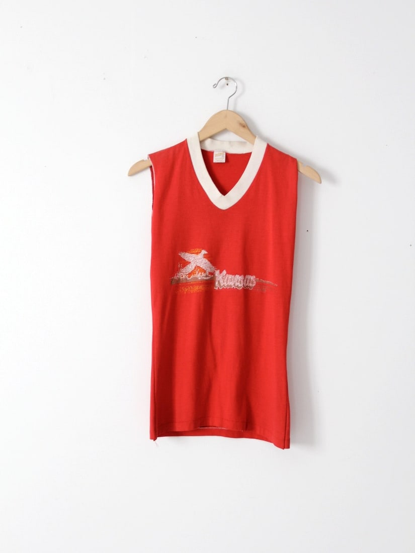1980s Vintage Kansas Red Tank Top with Bird Design: Embrace the vintage charm with this 1980s Kansas tank top. Featuring a vibrant red color, this piece is adorned with a white and gold screen print of a soaring bird and the word 'KANSAS' across the ch