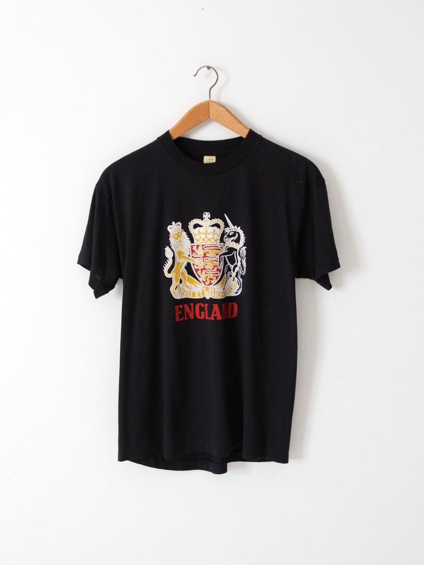 1980s Vintage England T-Shirt with Royal Coat of Arms: 1980s Vintage England T-Shirt with Royal Coat of Arms Showcase your British pride with this vintage 1980s England graphic t-shirt. The classic black crew neck features the Royal British Coat of Arms p