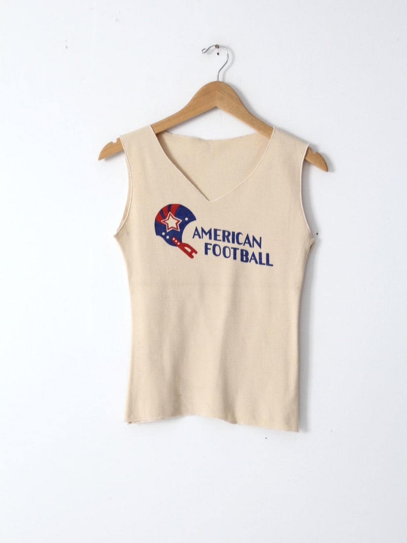Vintage American Football V-Neck Tank Top Cream Knit: Vintage American Football V-Neck Tank Top Cream Knit This vintage American Football tank top combines comfort and style with its cream knit fabric and bold red and blue graphic design. The fitted tank