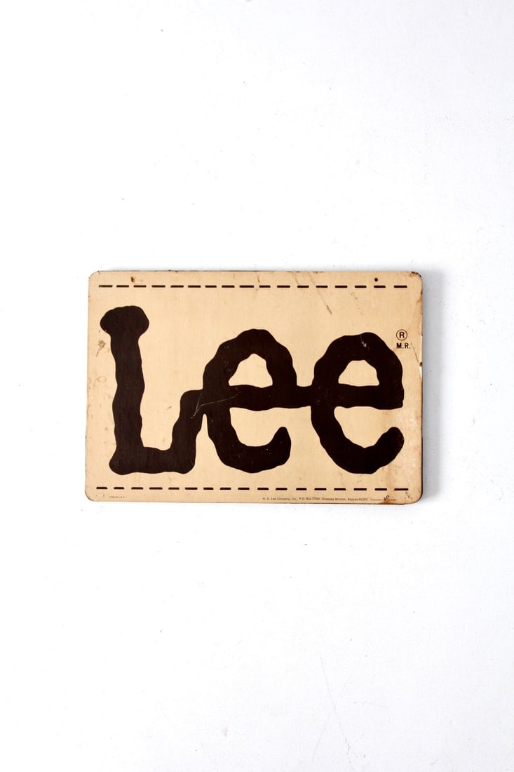 Vintage Lee Sign Mounted on Wood with Iconic Logo: Vintage Lee Sign Mounted on Wood with Iconic Logo This vintage Lee brand sign features the iconic Lee logo in tan and brown tones, beautifully mounted on a wooden backboard. The sign showcases a rusti